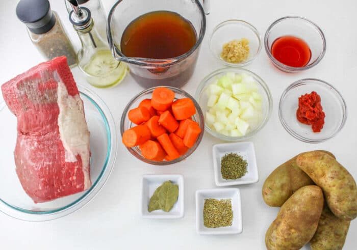 Ingredients for a classic beef stew: tender chunks of chuck roast, carrots, onions, and potatoes are braised in a rich broth with aromatic spices, tomato paste, garlic, and a splash of red wine all on a white surface.