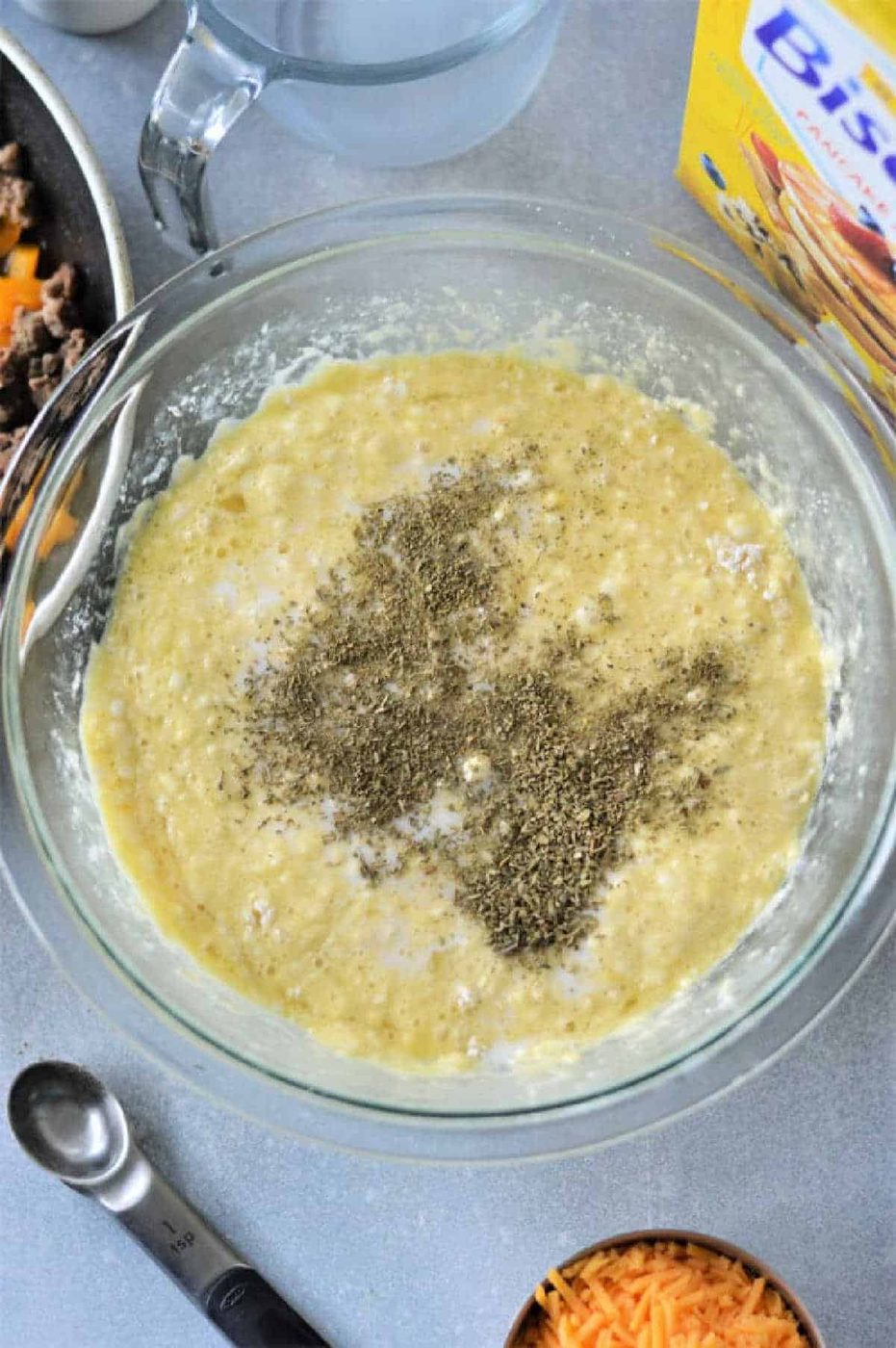 A glass bowl with batter and dried herbs, surrounded by cheese, Bisquick mix, and a measuring spoon—solid ingredients for making a good Breakfast Casserole with Bisquick.
