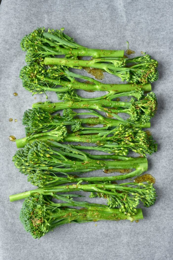 Fresh broccolini with seasoning drizzled on top, arranged on parchment paper, ready to be transformed into good oven roasted broccolini.
