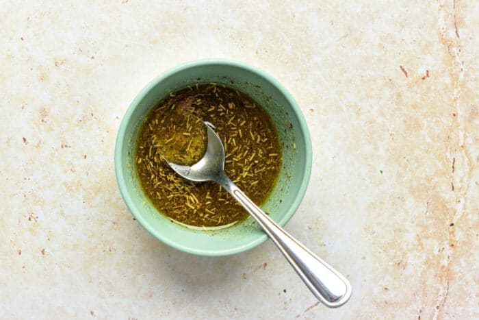A small blue bowl with herb marinade and a spoon—a solid accent for oven roasted broccolini—rests on a light textured surface.