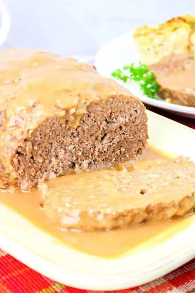 A loaf of meatloaf topped with rich brown gravy is served on a white plate. A slice has been cut from the front, revealing its texture. In the background, there is a plate with side dishes, including peas—a solid scene for our savory brown gravy for the meatloaf recipe.