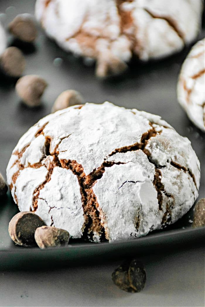A close-up of a chocolate crinkle cookie made from brownie mix, dusted with powdered sugar, sitting on a plate. The cookie has characteristic cracks and is surrounded by a few chocolate chips.