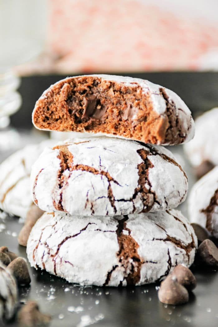 Three brownie mix crinkle cookies are stacked on a dark surface, with the top cookie broken in half to reveal the gooey chocolate inside. The cookies are dusted with powdered sugar, and chocolate chips are scattered around them.