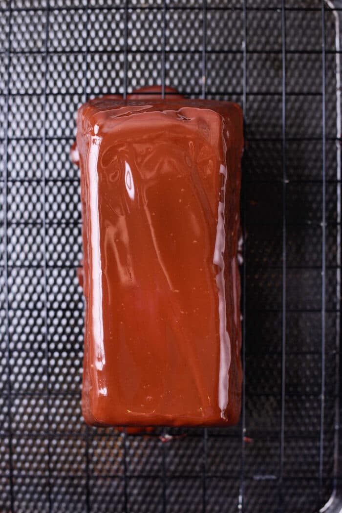 A rectangular brownie coated with a smooth, glossy layer of chocolate glaze rests on a black wire cooling rack, placed atop a textured black surface.