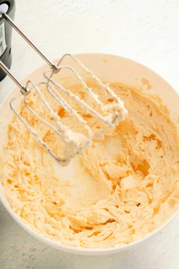 A mixing bowl filled with creamy, light yellow butter awaits. An electric mixer with two beaters, coated in the same velvety mixture for a luscious brownie frosting, rests above the bowl on a pristine white countertop.