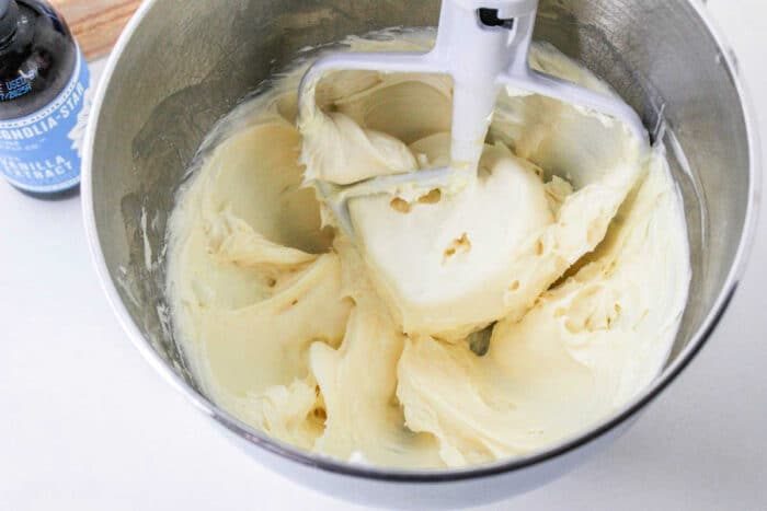 A stand mixer bowl filled with creamy, smooth batter. A paddle attachment is visible, partially coated with the batter. A vanilla extract bottle and a hint of brownie frosting are partially visible on the left side of the image.