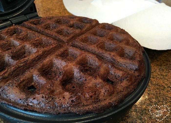 A good chocolate waffle, reminiscent of brownie waffles, sits enticingly inside a waffle maker, partially opened on the countertop.
