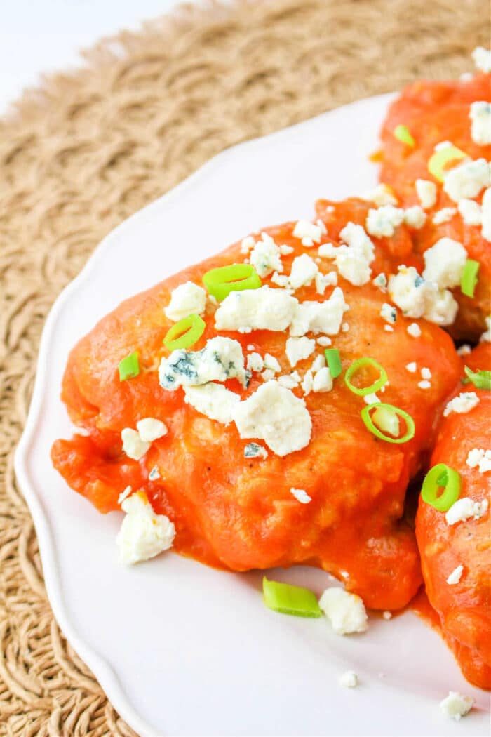 Buffalo chicken thighs are topped with blue cheese crumbles and sliced green onions, elegantly displayed on a white plate.