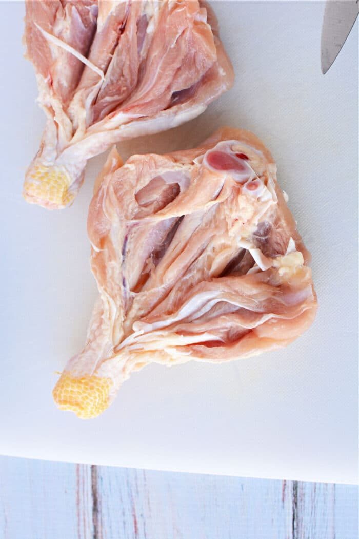 Two raw, butterflied chicken legs rest on a white cutting board, partially cut open, with a knife visible in the top right corner.