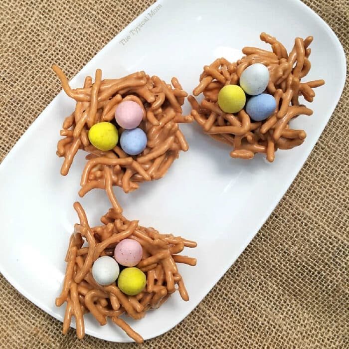 Three butterscotch haystacks shaped into cookie nests, made of pretzel sticks and filled with colorful candy eggs, sit on a white plate.