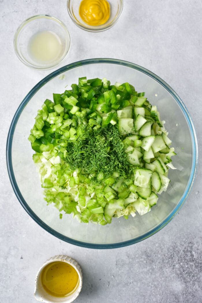 Bowl of chopped green vegetables and herbs solid for a refreshing cabbage salad, with small bowls of mustard, lemon juice, and dressing nearby.