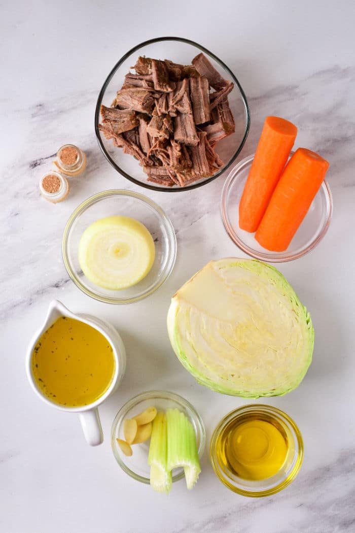 On a marble surface lie the ingredients for a good cabbage stew recipe: shredded beef, carrots, onion, cabbage, broth, garlic, celery, olive oil, and spices.
