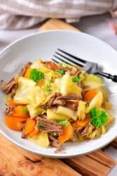 Plate of corned beef with a cabbage stew twist, complemented by carrots and garnished with parsley, all elegantly presented on a wooden surface.