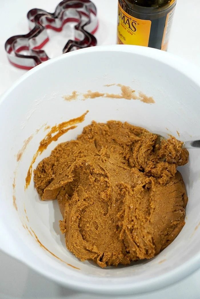A white bowl contains rich gingerbread dough with a spoon sticking out, hinting at good gingerbread cookies to come. A bottle and cookie cutters are thoughtfully placed in the background, ready for action.