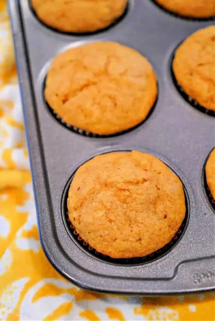 A muffin tin holds six freshly baked golden brown Cake Mix Pumpkin Banana Muffins on a yellow patterned cloth.