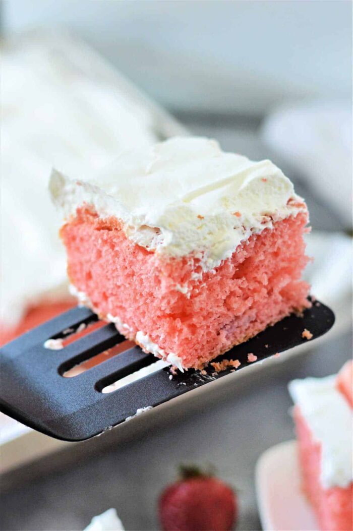 A slice of pink Strawberry Poke Cake Mix with Fresh Strawberries and white frosting rests on a spatula, with a strawberry visible in the background.
