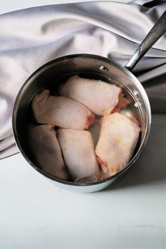 frozen chicken thighs rest in a metal pot filled with water on a marble surface, set against a silver cloth backdrop—a solid start for those wondering, Can you boil frozen chicken?.