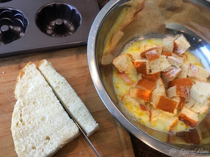 Sliced bread rests on a cutting board while diced pieces soak in a metal bowl, absorbing flavors for a good caramel apple bread pudding. A baking mold awaits in the background, ready for the final creation.