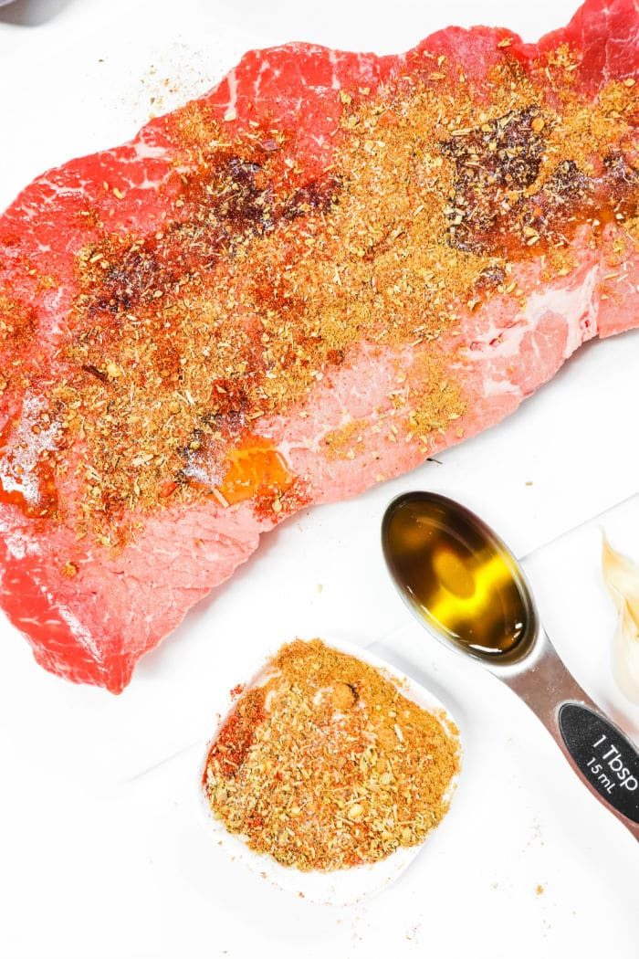 Raw marinated flank steak with seasoning, a tablespoon of oil, and a garlic clove on a white surface—solid for slow-cooking into tender carne asada in your crock pot.