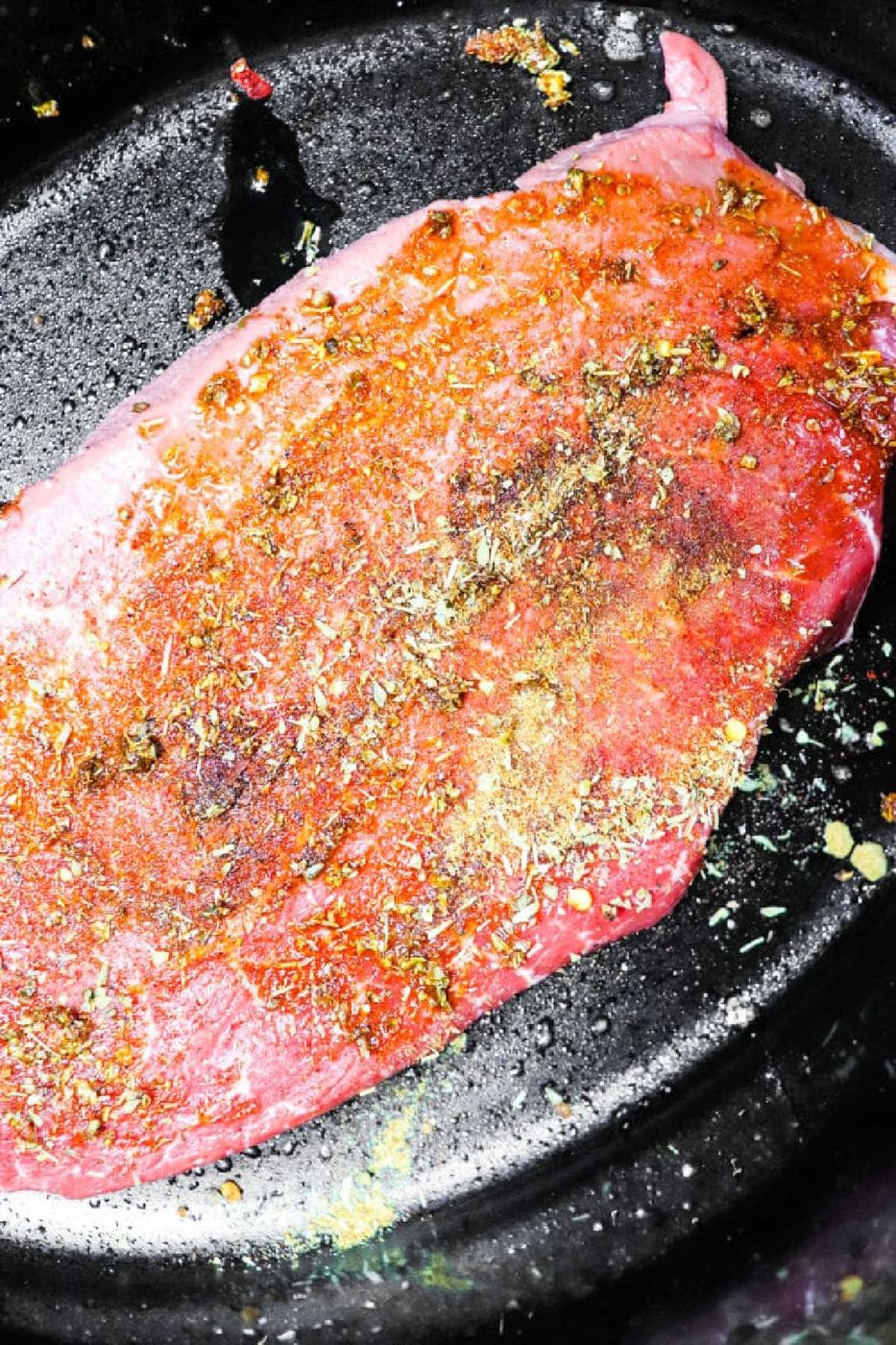 Raw seasoned beef steak on a black surface, sprinkled with various spices and herbs, solid for making Carne Asada Crock Pot recipes.