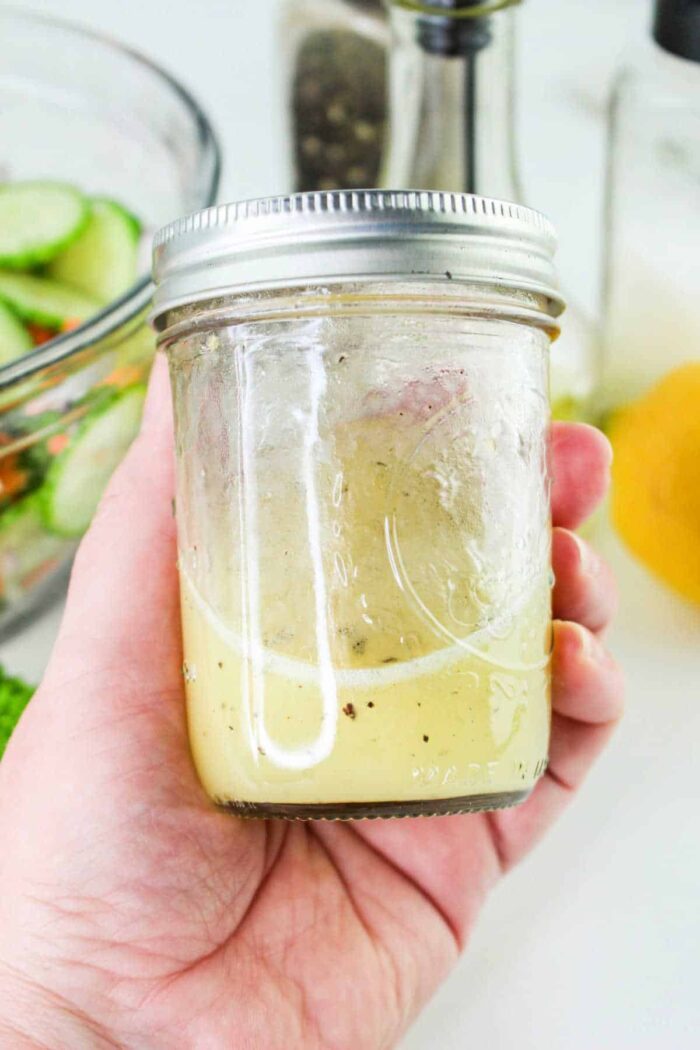 A hand is holding a glass jar filled halfway with light-colored salad dressing, solid for a refreshing Carrot and Cucumber Salad. The jar has a metal screw-top lid. In the background, there is a bowl of sliced cucumbers, a pepper grinder, a bottle, and a lemon.