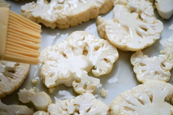 A brush spreads oil on sliced cauliflower pieces arranged on a baking sheet, preparing them for good cauliflower steak air fryer cooking.