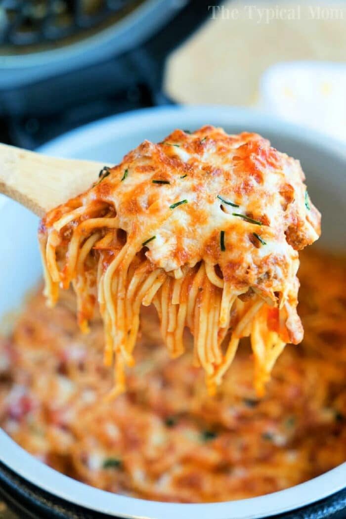 A wooden spoon lifting cheesy Ninja Foodi spaghetti from a pot, with melted cheese and herbs on top.