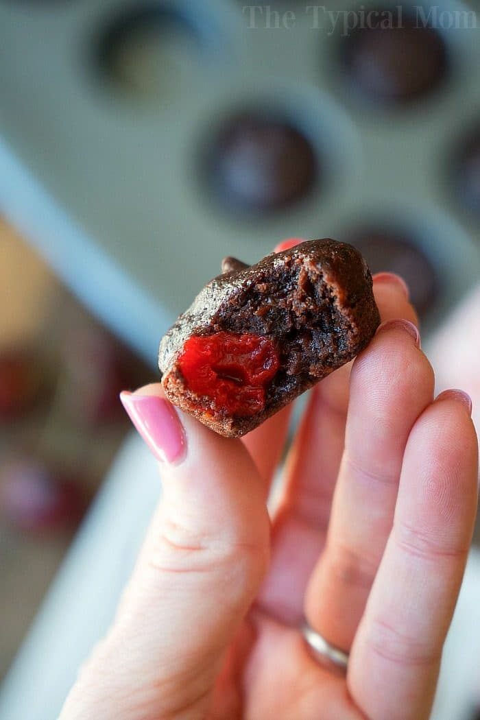 A close-up of a hand holding a small chocolate dessert with a bite taken out, revealing a bright red cherry filling inside. The background shows a blurred muffin tray filled with these good cherry brownies.