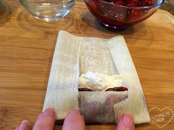 A person skillfully folds a egg roll wrapper filled with creamy cherry cheesecake and fruit filling on a wooden board.