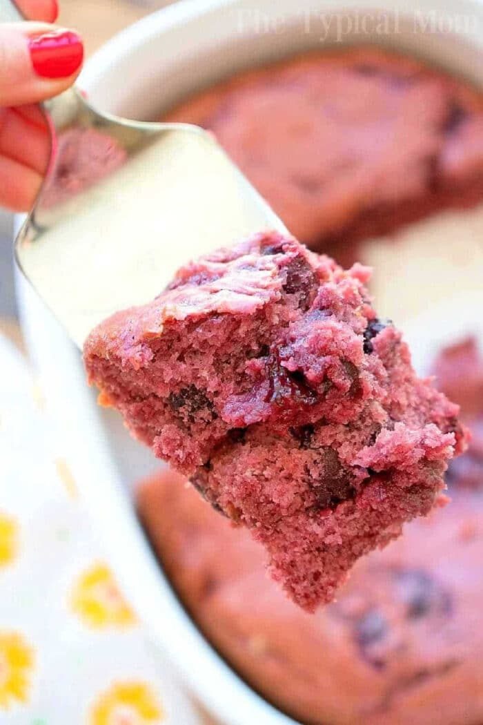 A close-up of a spatula holding a piece of cherry pie filling dump cake above a baking dish.