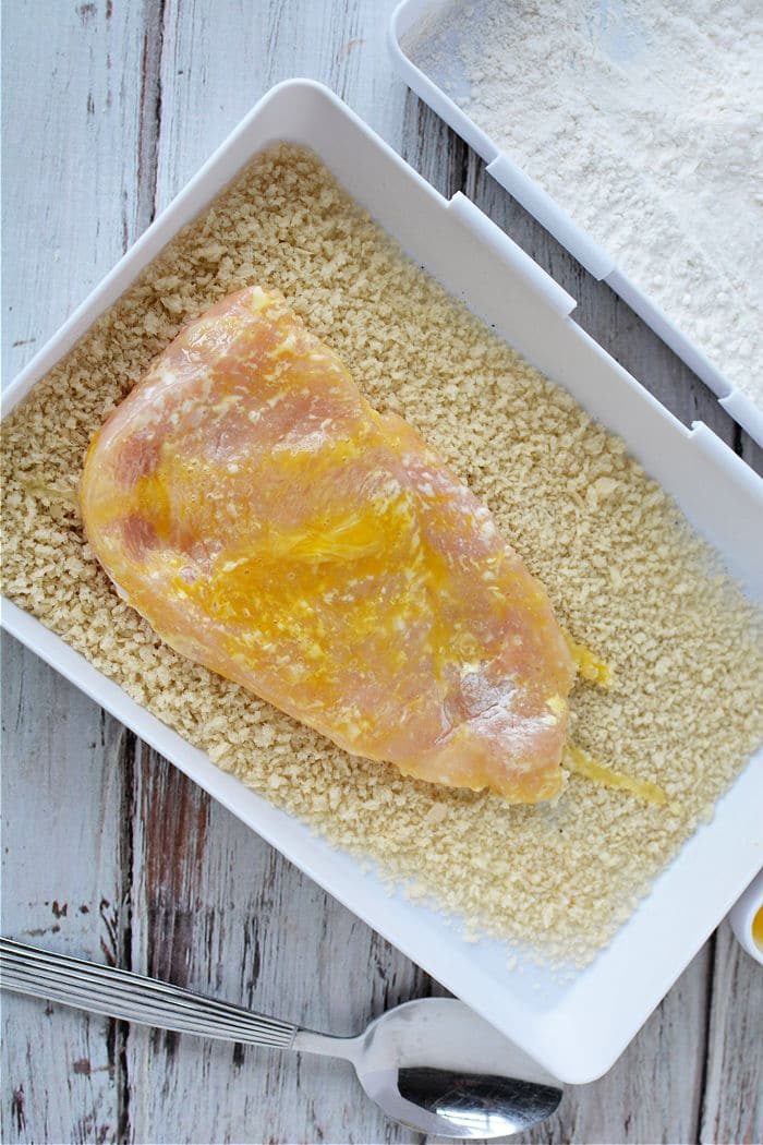 Raw chicken cutlet coated in egg on a tray of breadcrumbs, ready to become good chicken cutlets in the air fryer, with a spoon nearby.