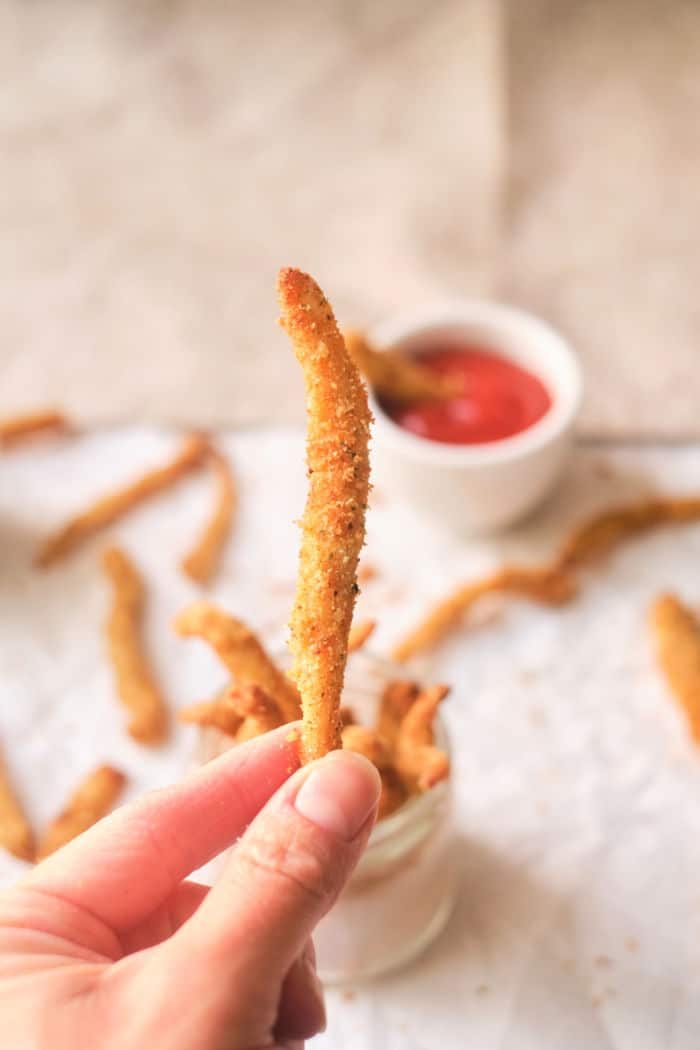 Close-up of a hand holding a crispy chicken fry, with more fries and dipping sauce in the background, reminiscent of chicken fries cooked to perfection in an air fryer.