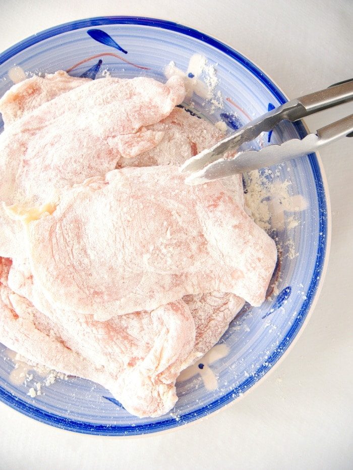 Raw chicken cutlet pieces, solid for creating a quick chicken marsala, coated in flour on a blue plate with metal tongs.