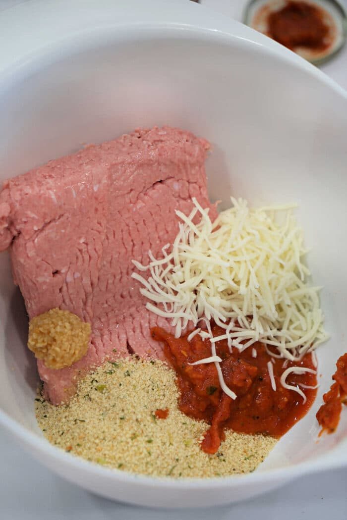Ground meat, shredded cheese, breadcrumbs, minced garlic, and tomato sauce in a white mixing bowl—solid for preparing a flavorful Chicken Parmesan Meatloaf.