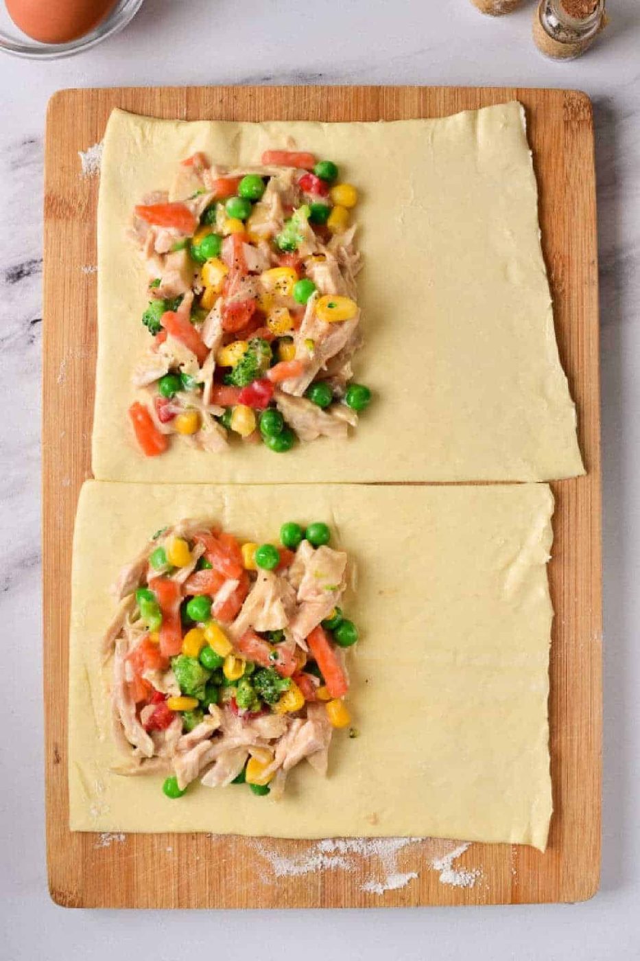 Two chicken pot pie puff pastry rectangles rest on a wooden board, each topped with a savory mix of vegetables and shredded chicken filling.