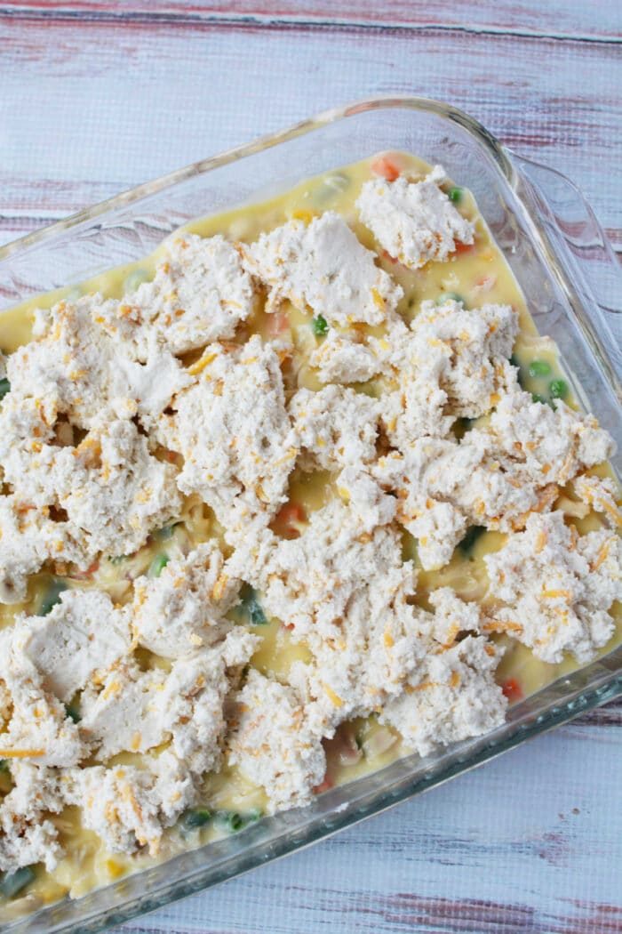 Unbaked chicken pot pie casserole topped with biscuits sits in a glass dish on a white wooden surface.