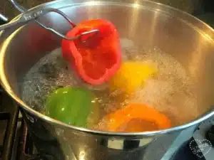 A stainless steel pot filled with boiling water on a stove holds green, yellow, and orange bell peppers. A metal utensil expertly demonstrates how to blanch bell peppers by submerging a red one into the water.