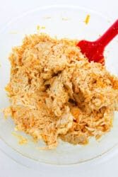 A glass bowl with shredded buffalo chicken dip being mixed by a red spatula, solid for topping your chicken stuffed bell peppers.