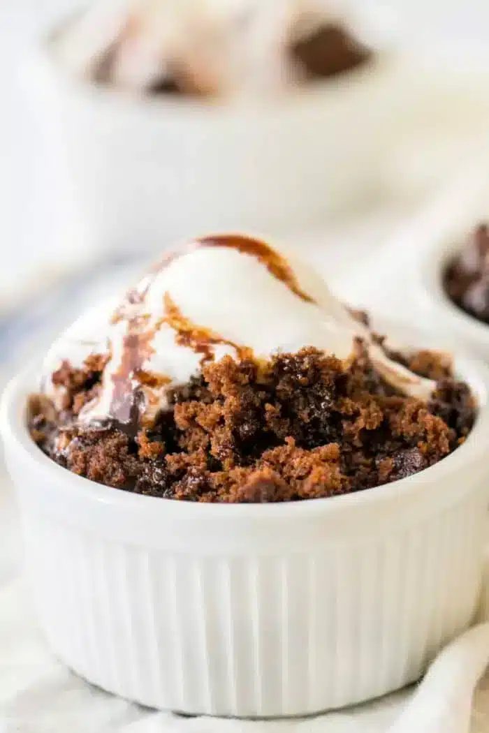 A white ramekin filled with a delectable hot water cake topped with a scoop of vanilla ice cream and drizzled with rich chocolate syrup. Another ramekin with a similar treat is visible in the blurred background.