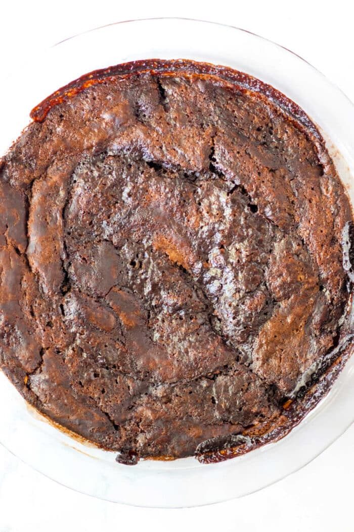 Aerial view of a round, baked chocolate cake in a clear dish on a white surface.