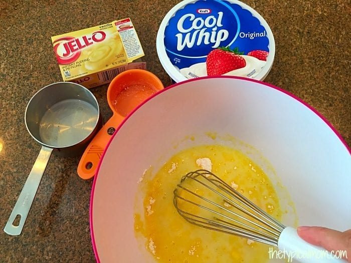 A bowl containing a yellow mixture being whisked. Surrounding the bowl are a box of instant lemon pudding mix, a tub of Cool Whip, a measuring cup, and an orange measuring spoon with some mix in it. A hand is holding the whisk, preparing what looks like tasty mug cake frosting.