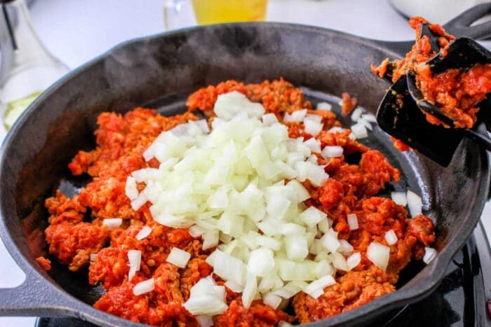 Chopped onions on top of raw ground meat in a black skillet, being stirred with a spatula—solid as the base for a flavorful chorizo stuffing or your favorite stuffing recipe.