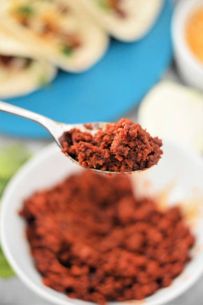 Close-up of a spoonful of seasoned chorizo over a bowl, hinting at the good flavors of a chorizo taco recipe, with tacos blurred in the background.