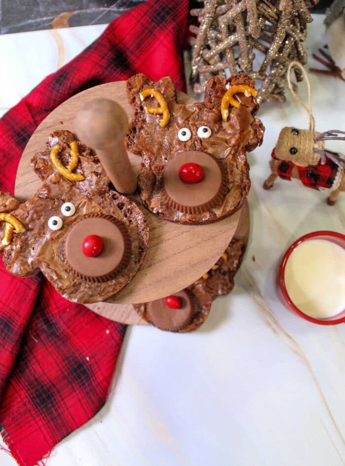 Reindeer-themed brownies with pretzel antlers and candy eyes sit alongside Christmas brownies on a wooden stand, next to a red plaid cloth and a drink.