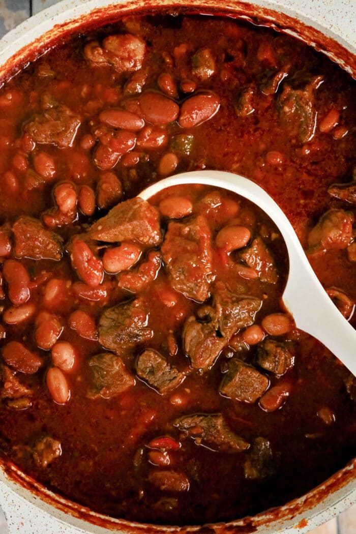 A white ladle scoops rich Chuck Roast Chili, brimming with beef and beans, from a pot filled with thick, red, hearty stew.