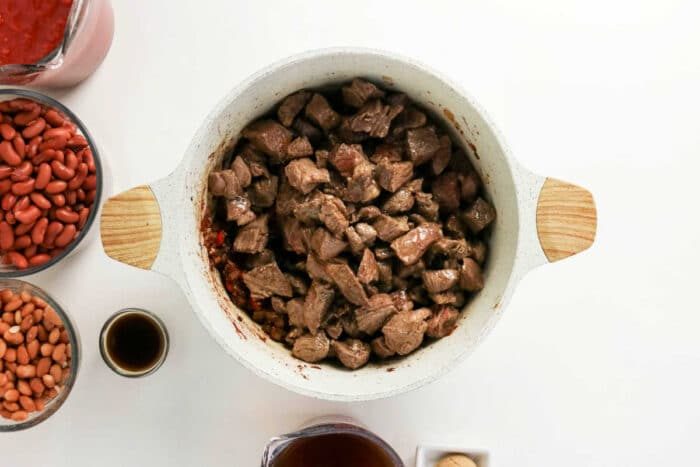 Overhead view of cubed beef in a white pot, solid for making Chuck Roast Chili, surrounded by beans, sauce, and seasonings on a white surface.