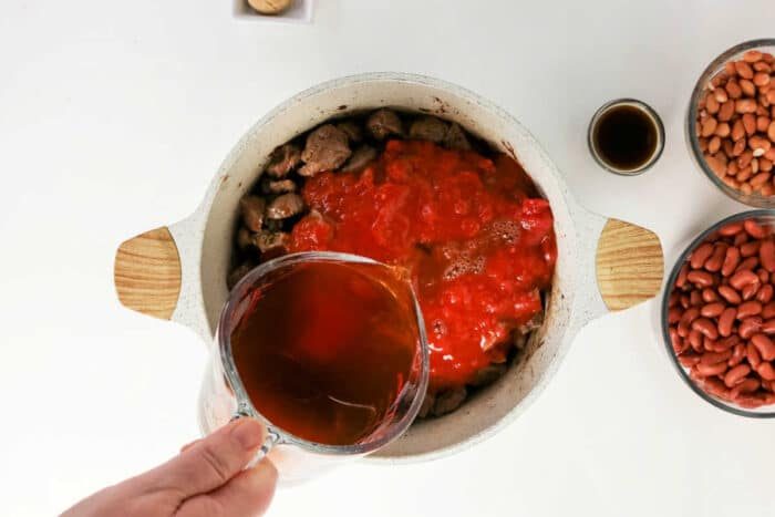 A hand pours broth into a pot with beef and tomatoes, surrounded by beans and sauce on a white table, creating the solid base for a hearty beef chili recipe.