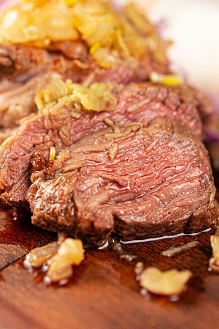 Close-up of juicy, sliced chuck roll steak topped with onions on a wooden cutting board.