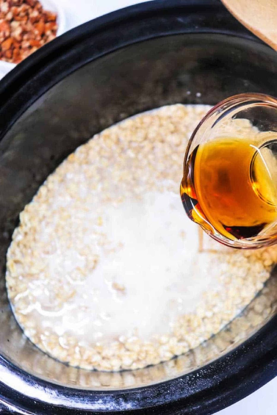 A hand pours a golden liquid into a slow cooker filled with oats and milk, preparing good Cinnamon Oatmeal.
