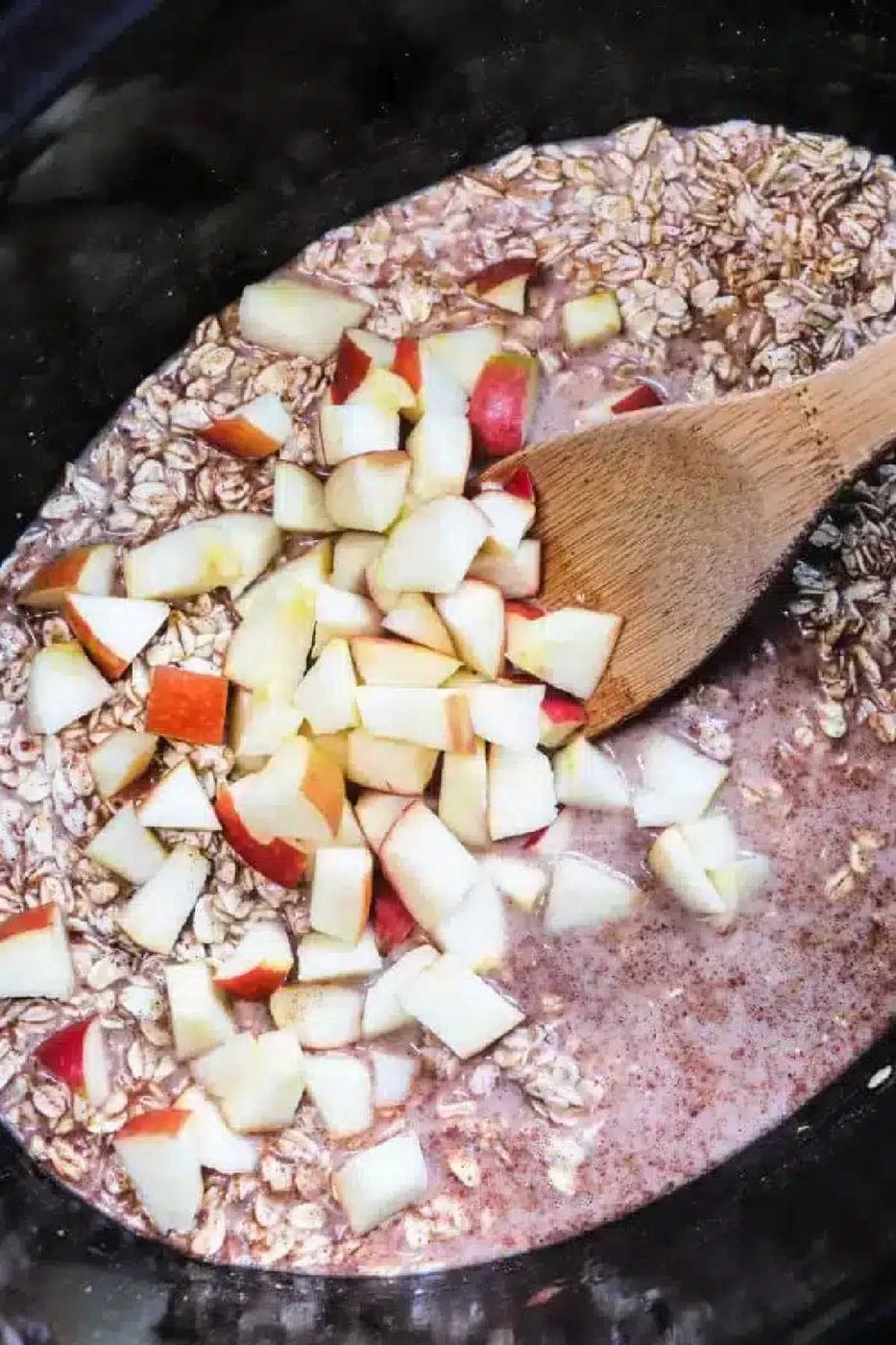 Wooden spoon mixing chopped apples, oats, and liquid in a black bowl or slow cooker for good cinnamon oatmeal.